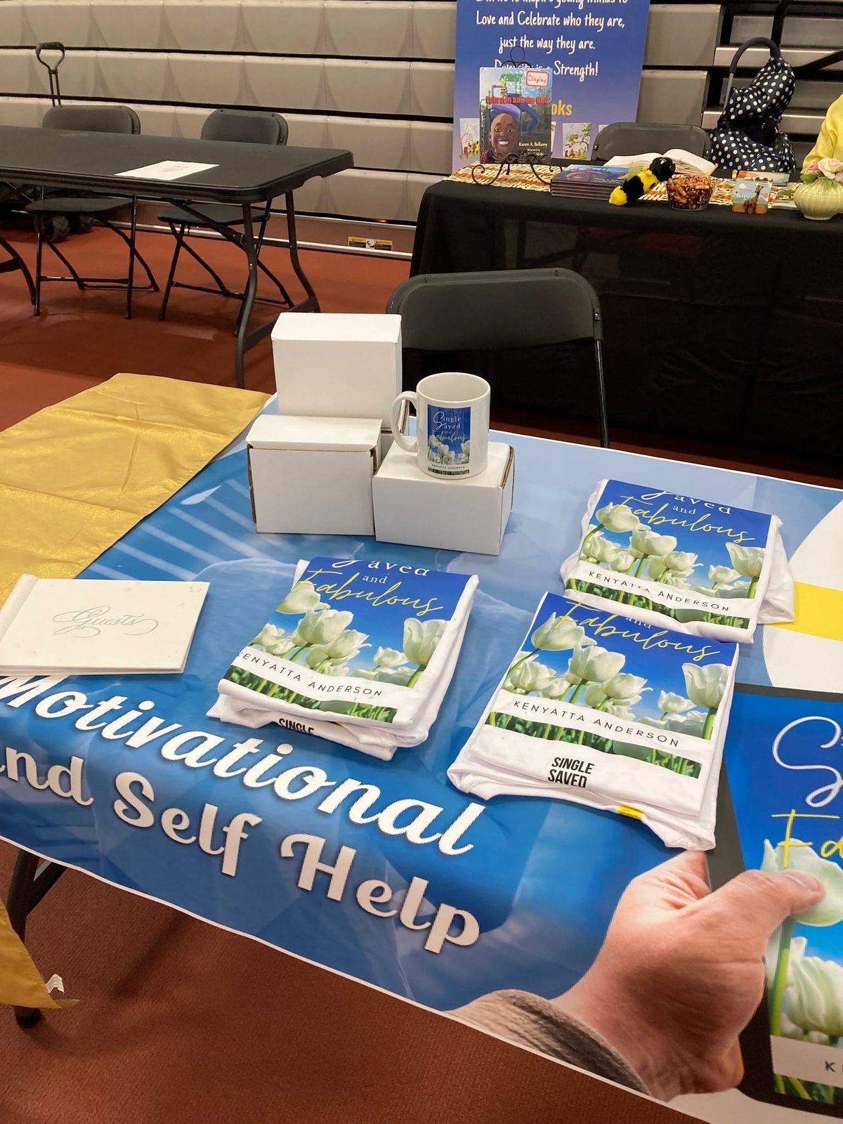 Book display table with motivational and self-help books, white boxes, blue banner, and promotional materials at an indoor event.
